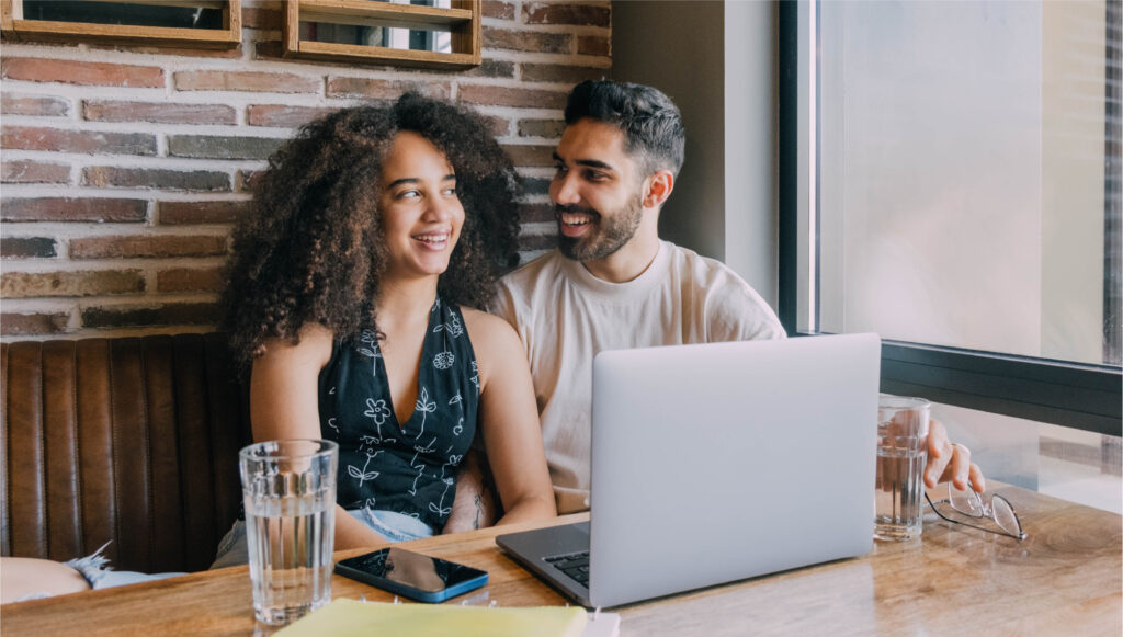 A smiling couple is sitting at a cafe table with an open laptop in front of them